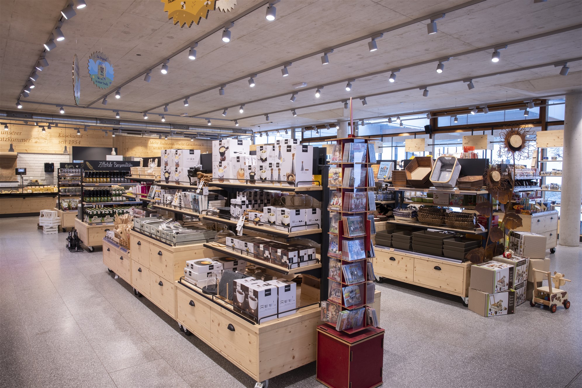 Large sales area with wooden shelves and many products. Bright ceiling lighting, wide aisles, household goods and food neatly presented.