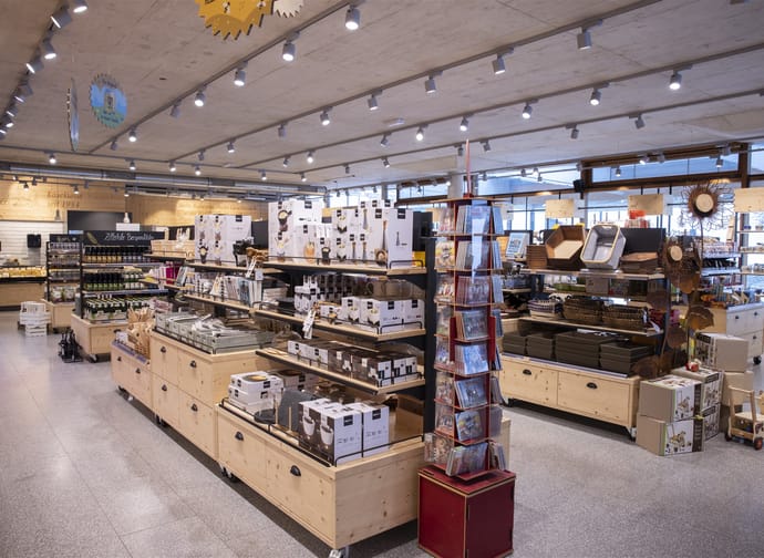 Large sales area with wooden shelves and many products. Bright ceiling lighting, wide aisles, household goods and food neatly presented.