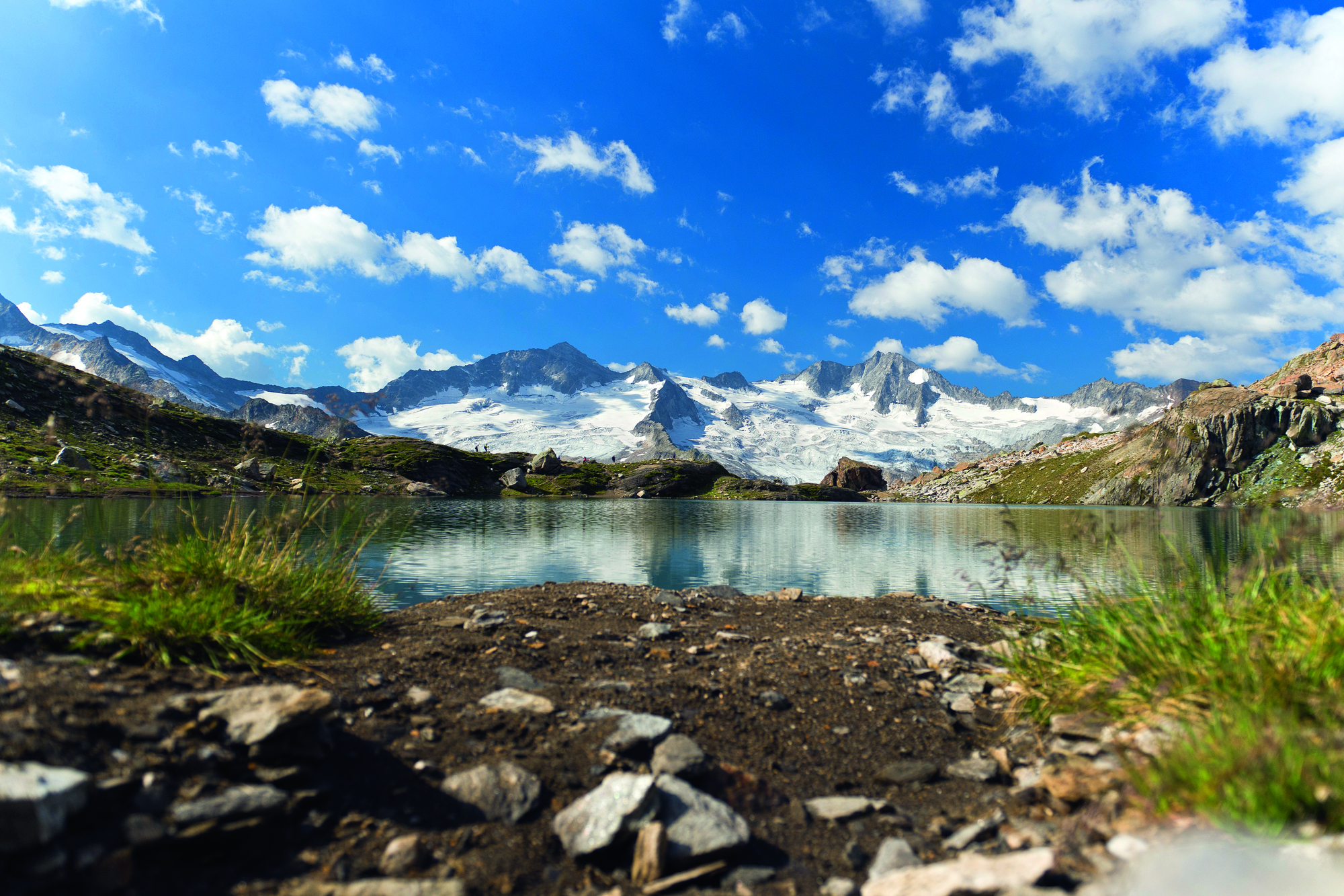Alpine mountain lake in Zillertal, Tyrol reflecting snow-covered peaks under a blue sky with white clouds, surrounded by green meadows and rocks.