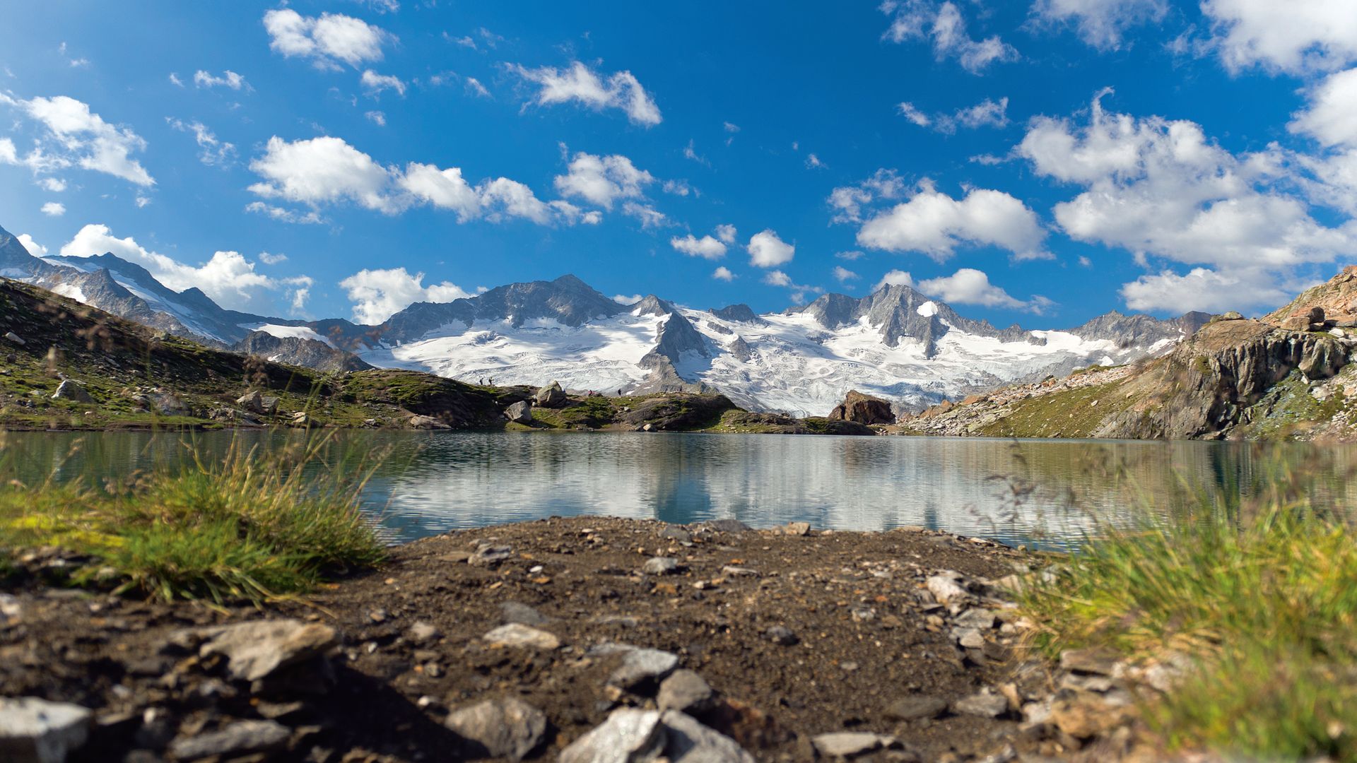 Alpine mountain lake in Zillertal, Tyrol reflecting snow-covered peaks under a blue sky with white clouds, surrounded by green meadows and rocks.
