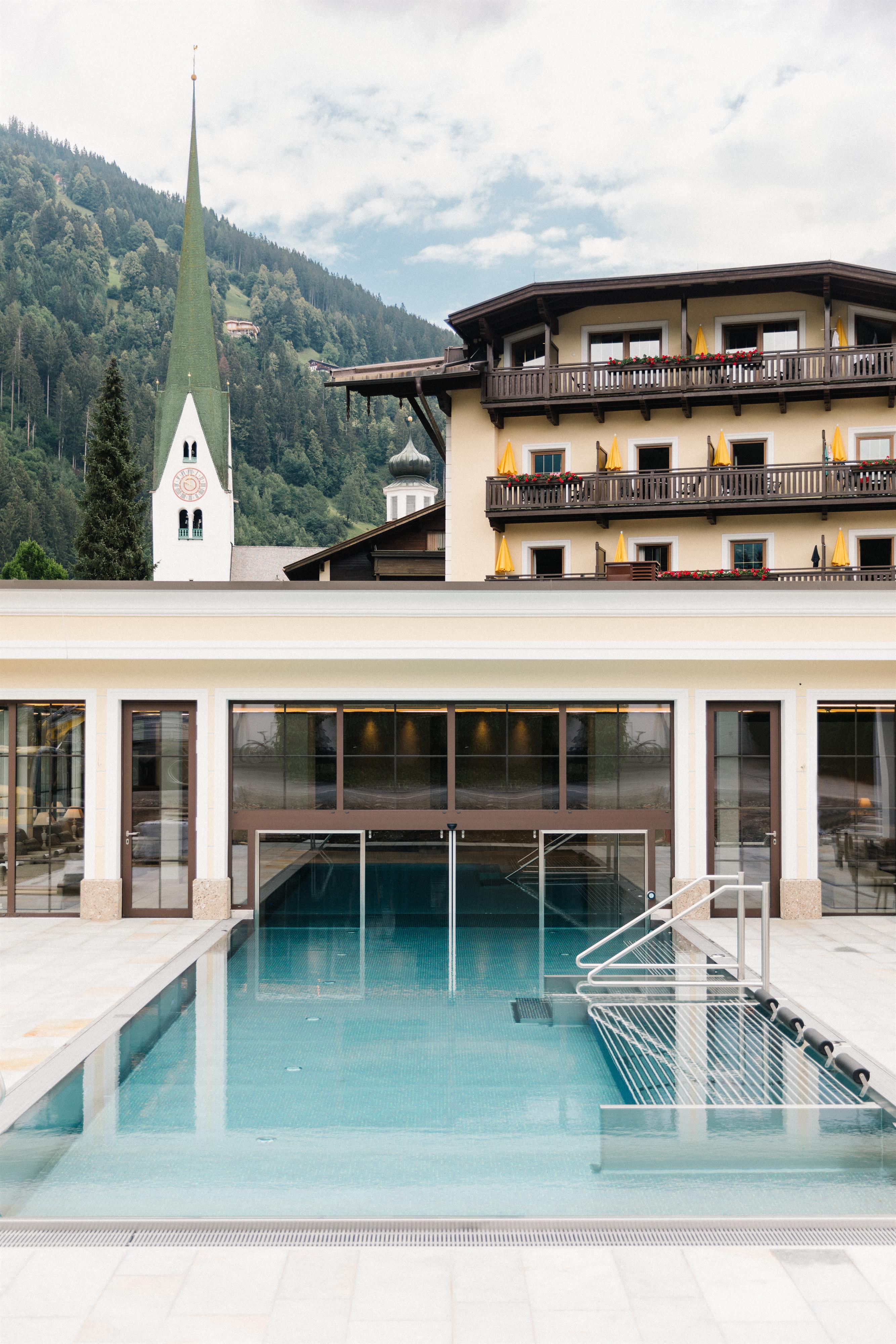 Innenansicht eines Schwimmbads mit großen Fenstern, Pool und Liegestühlen, im Hintergrund ein Gebäude mit Balkon.