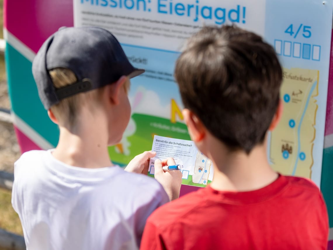 Mission Egg Hunt Mayrhofen-Hippach – Riddle at the Giant Easter Egg | Tyrol Two children stand in front of a giant Easter egg from the “Mission Egg Hunt” in Mayrhofen-Hippach, reading the riddle displayed on it.