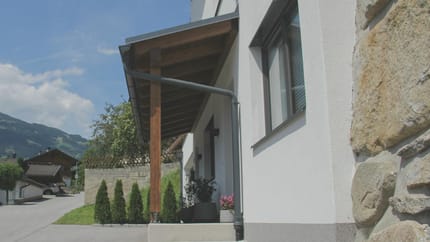 Weiße Hausfassade mit Fenster, Holzüberdachung und Steinmauer im Vordergrund, bei schönem Himmel.