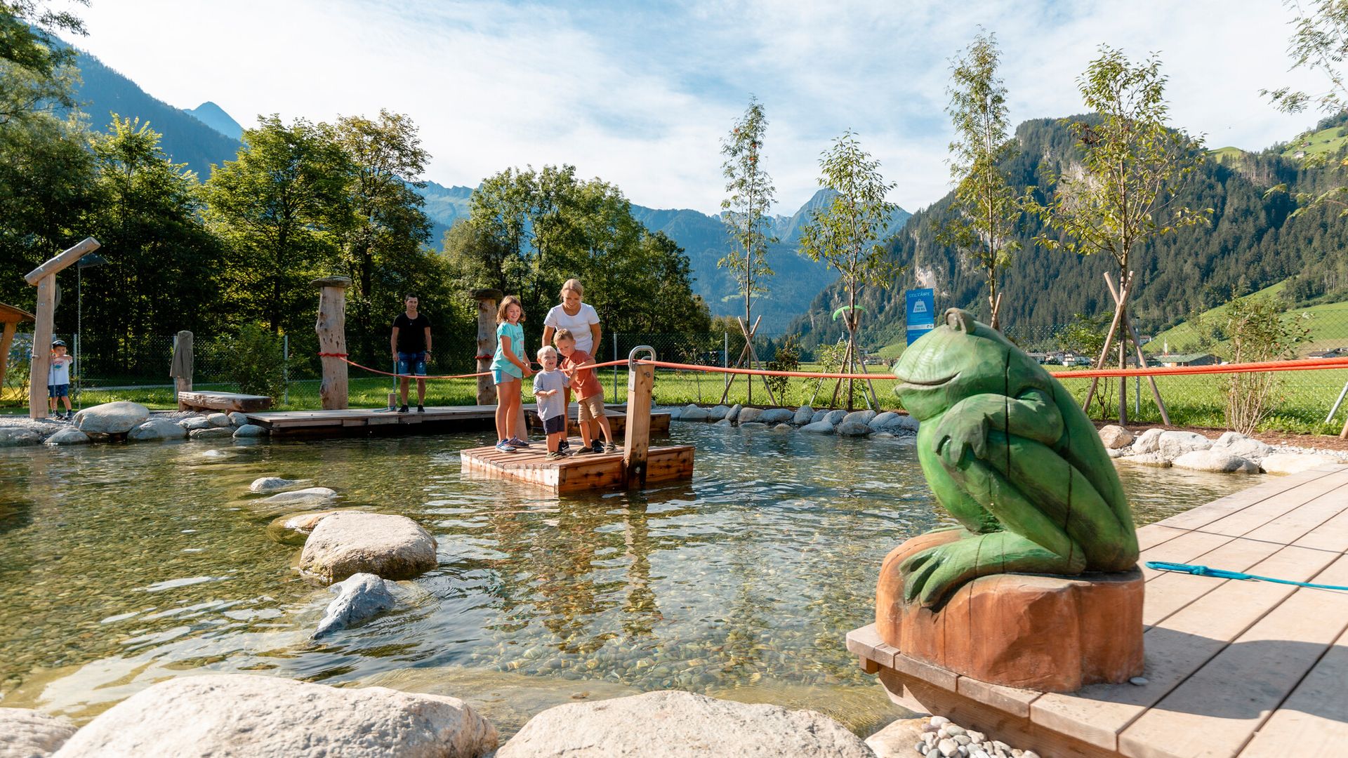 Familie auf einem Holzfloss im flachen Wasser des Spielplatzes Auenland Sidan in Mayrhofen-Hippach. Im Vordergrund eine geschnitzte Froschfigur, im Hintergrund Berge, Bäume und blauer Himmel.