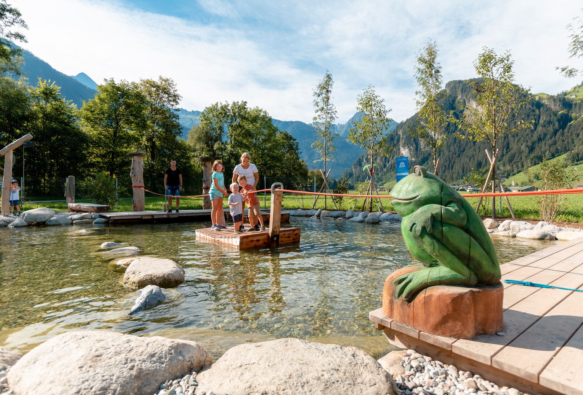 Family on a wooden raft in the shallow water of the Auenland Sidan playground in Mayrhofen-Hippach. In the foreground, a carved frog figure; in the background, mountains, trees, and blue sky.