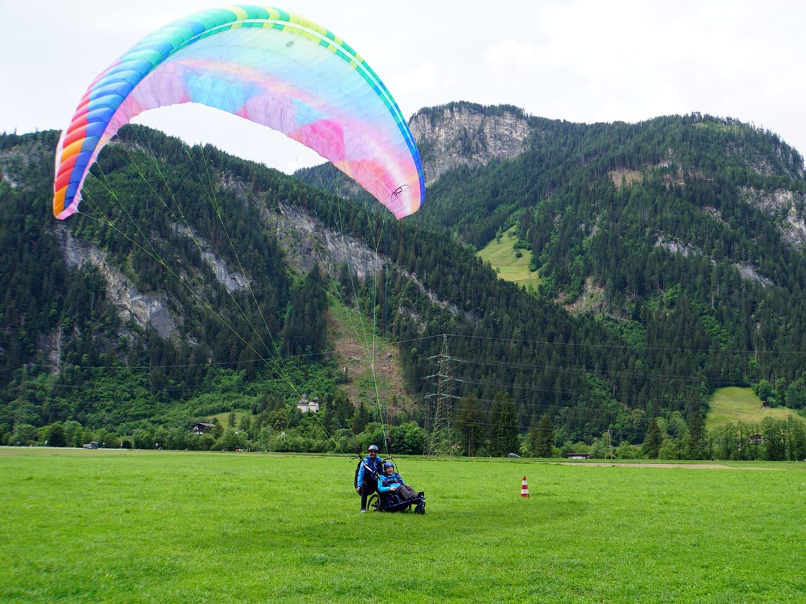 Rollstuhlgestütztes Gleitschirmfliegen - Landung Ein Pilot landet mit einem rollstuhlgestützten Gleitschirm auf einer grünen Wiese in Mayrhofen, Zillertal. Der bunte Schirm schwebt noch in der Luft, während bewaldete Berge die Kulisse bilden.