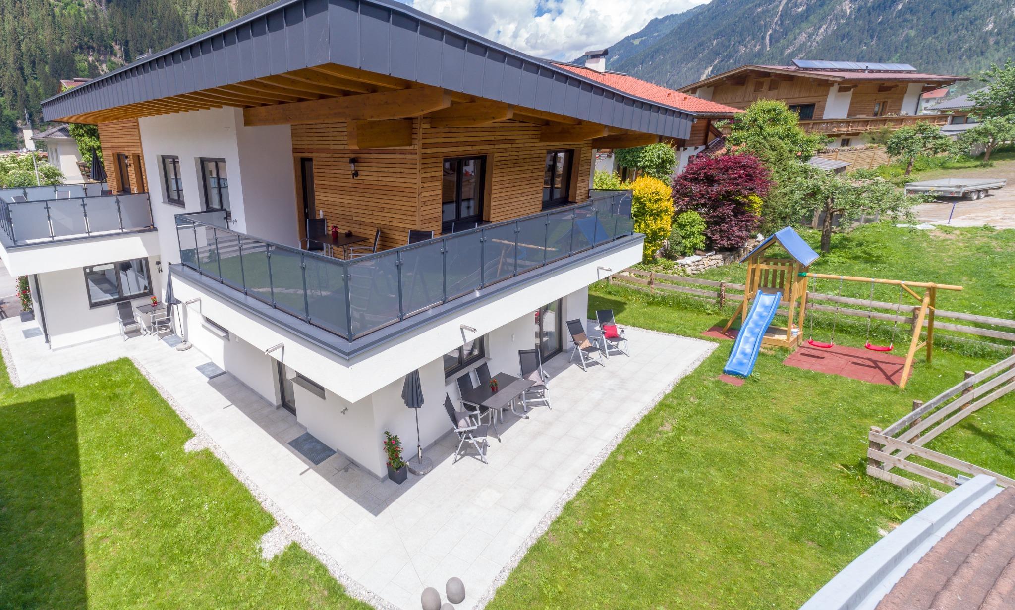 Modern house with balcony, garden, and playground, surrounded by mountains and trees.