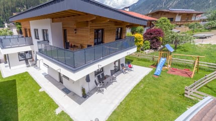 Modern house with balcony, garden, and playground, surrounded by mountains and trees.