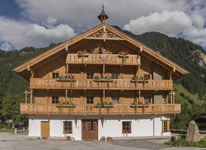 Traditionelles Holzhaus mit drei Etagen, Balkonen mit Blumen, vor grünen Bergen und Wolken am Himmel.