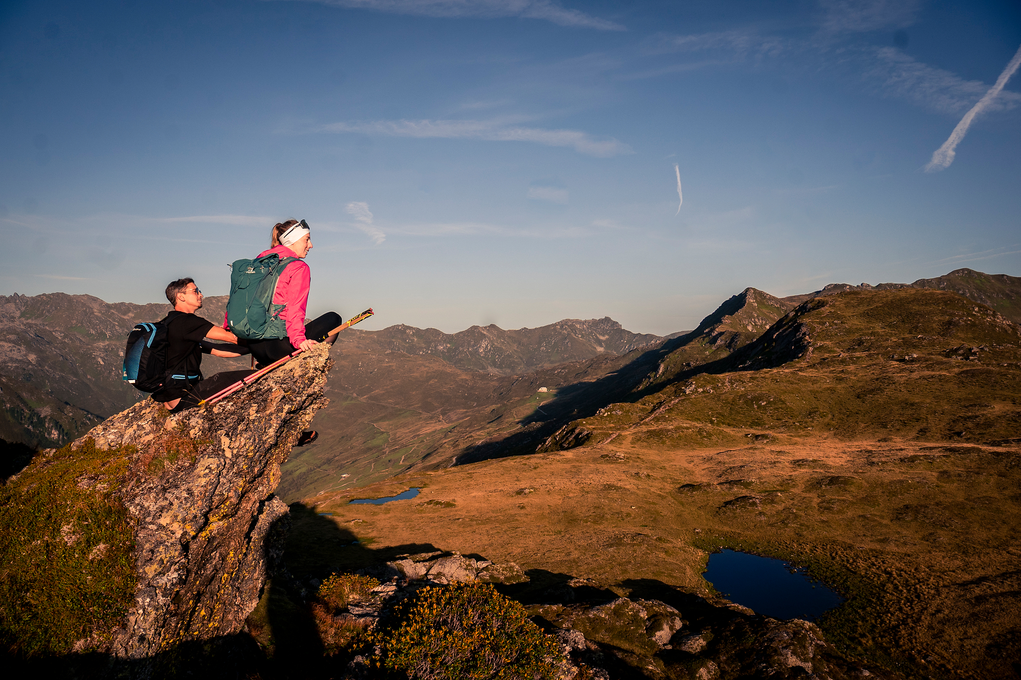 Two hikers sit on a rocky outcrop at sunrise with a sweeping view over the rolling hills and mountain peaks of the Zillertal - surrounded by a golden morning atmosphere and a clear view into the distance.