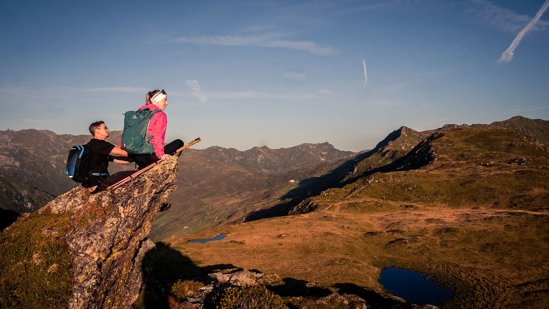 Zwei Wanderer sitzen bei Sonnenaufgang auf einem Felsvorsprung mit weitem Blick über die sanften Hügel und Berggipfel des Zillertals – umgeben von goldener Morgenstimmung und klarer Fernsicht.