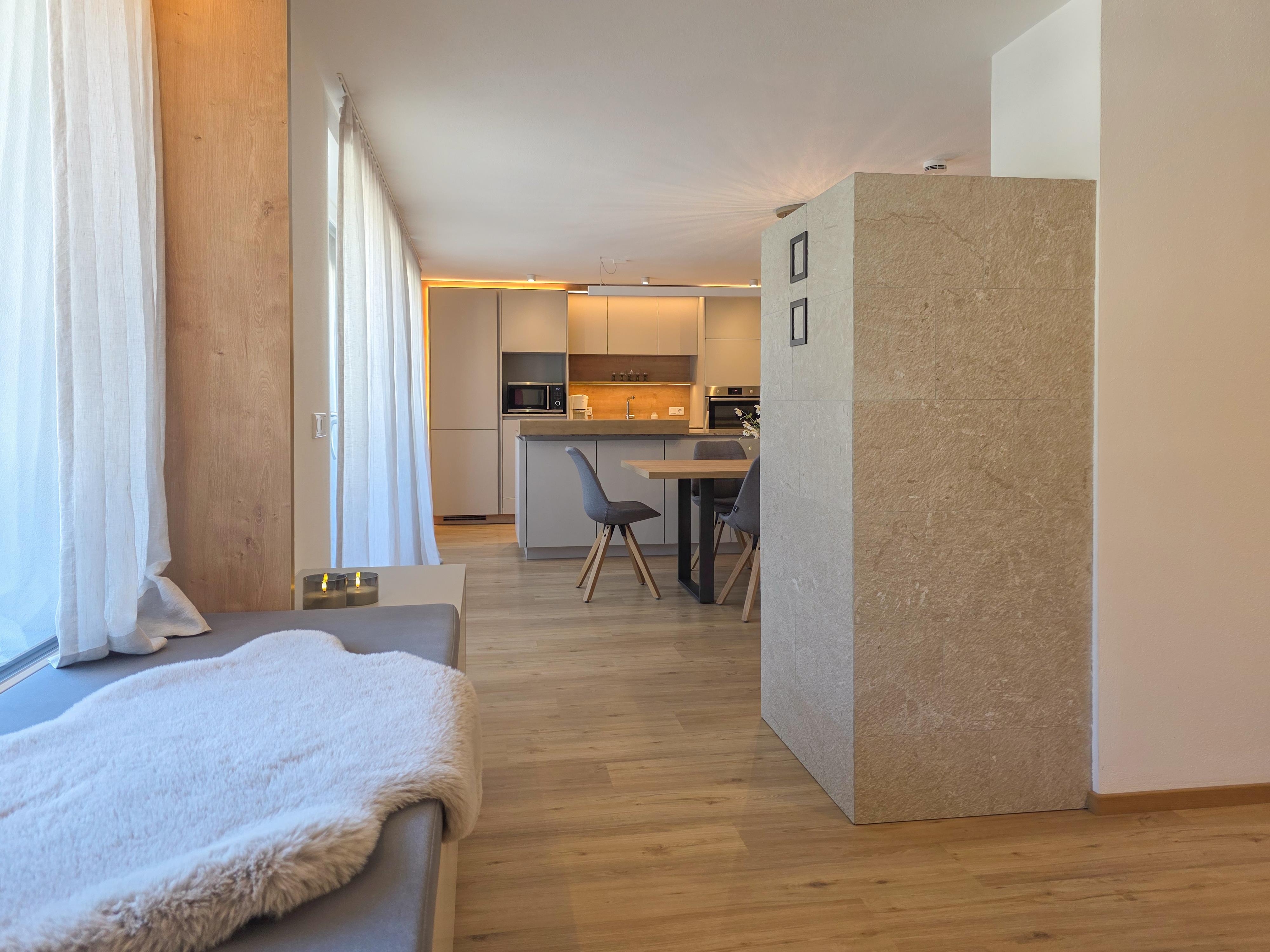 Gemütliches Wohnzimmer mit Sofa, Felldecke, großen Fenstern und offener Küche im Hintergrund.