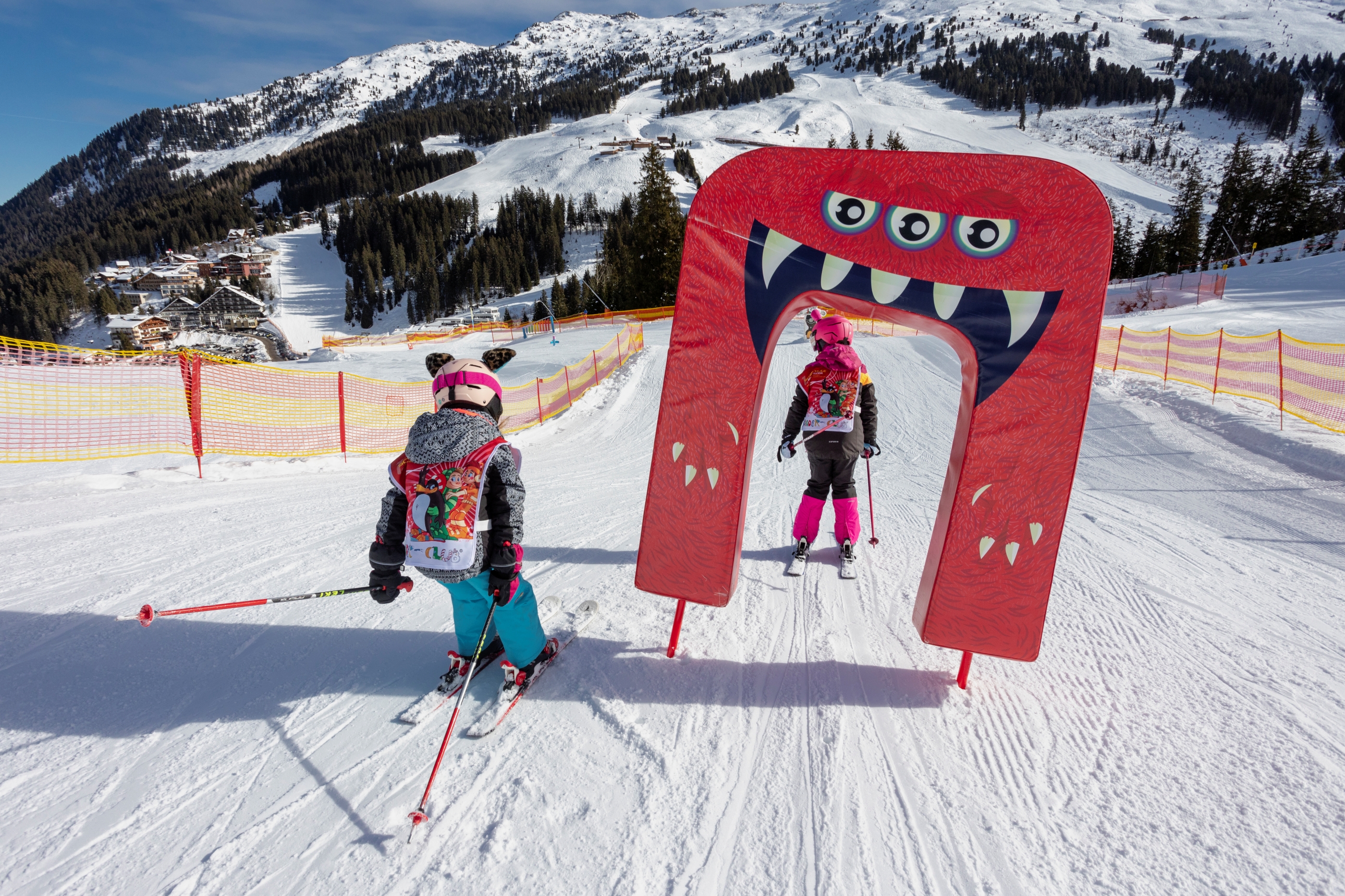 Zwei Kinder fahren auf Ski durch die Kidsarea in Hochfügen mit Blick auf Hotels, Lifte und das Bergpanorama im Hintergrund.
