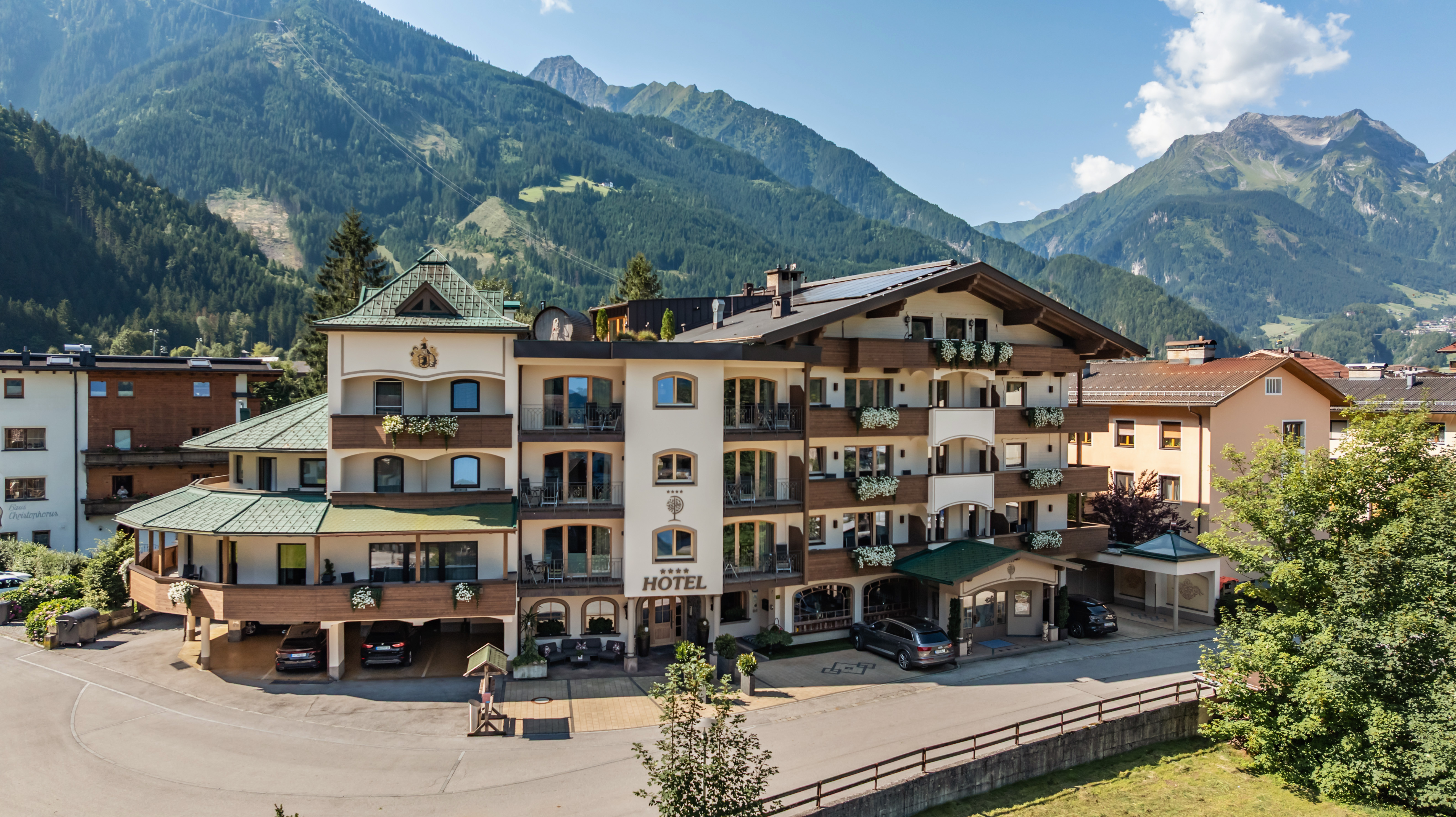 The image shows a large, multi-story hotel building in Alpine style, surrounded by an impressive mountain landscape. The hotel has bright facades with wooden balconies, adorned with flower boxes. Cars are parked in front of the entrance.