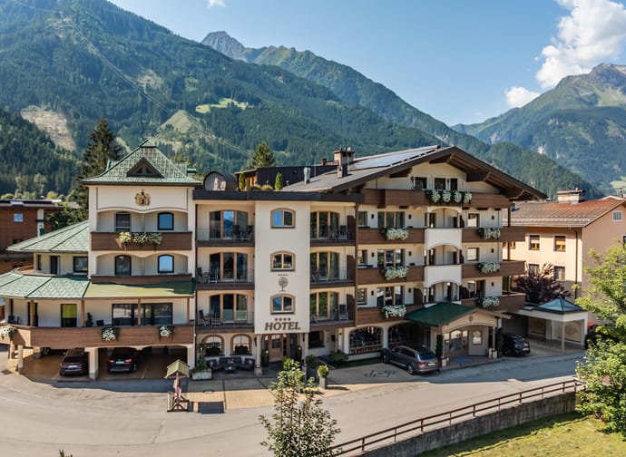 The image shows a large, multi-story hotel building in Alpine style, surrounded by an impressive mountain landscape. The hotel has bright facades with wooden balconies, adorned with flower boxes. Cars are parked in front of the entrance.