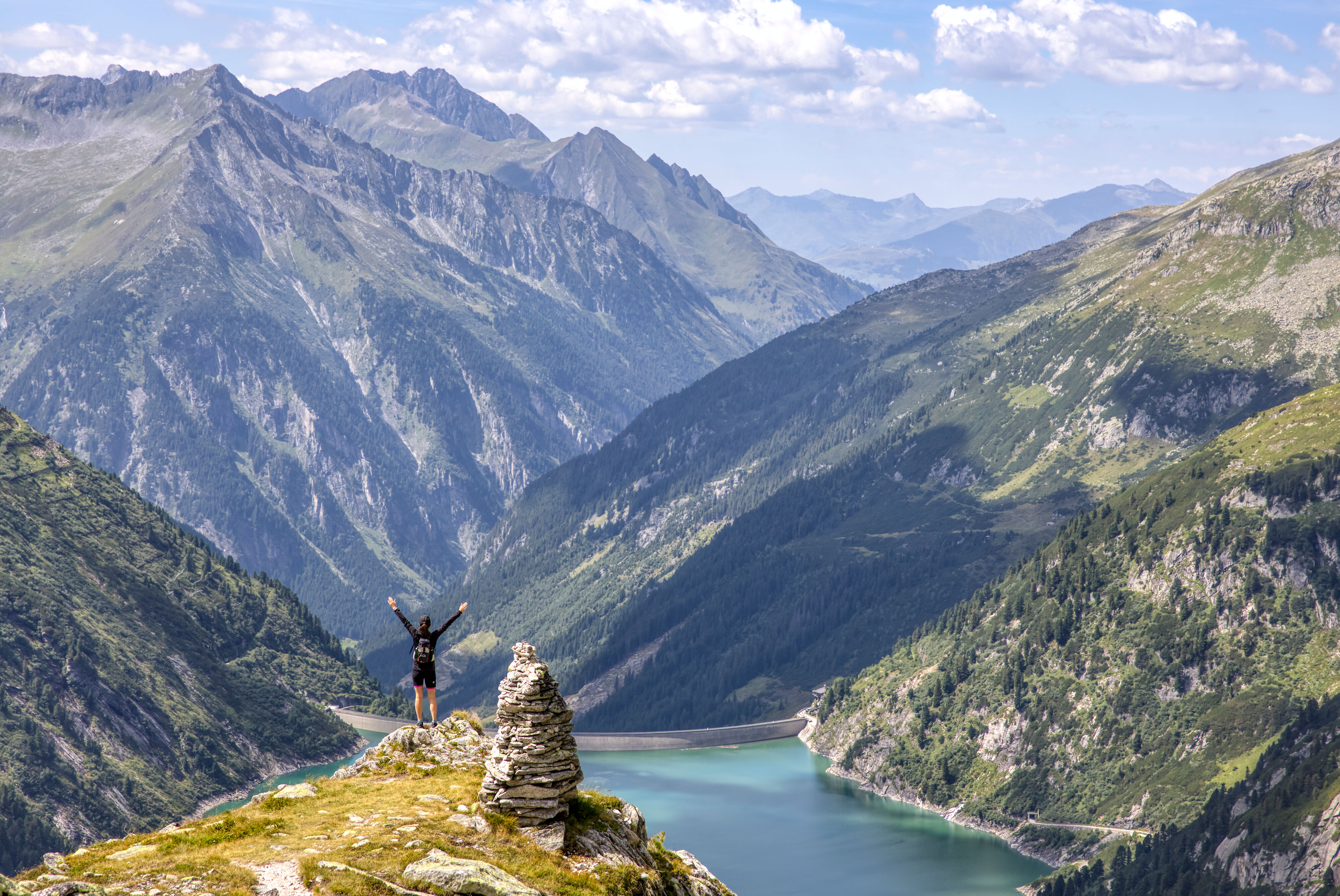 Eine Person steht auf einem Felsen hoch über dem Zillergründl-Speicher im Zillergrund. Der Blick reicht über das türkisfarbene Wasser, steile Berghänge und schroffe Gipfel bis zum Horizont.