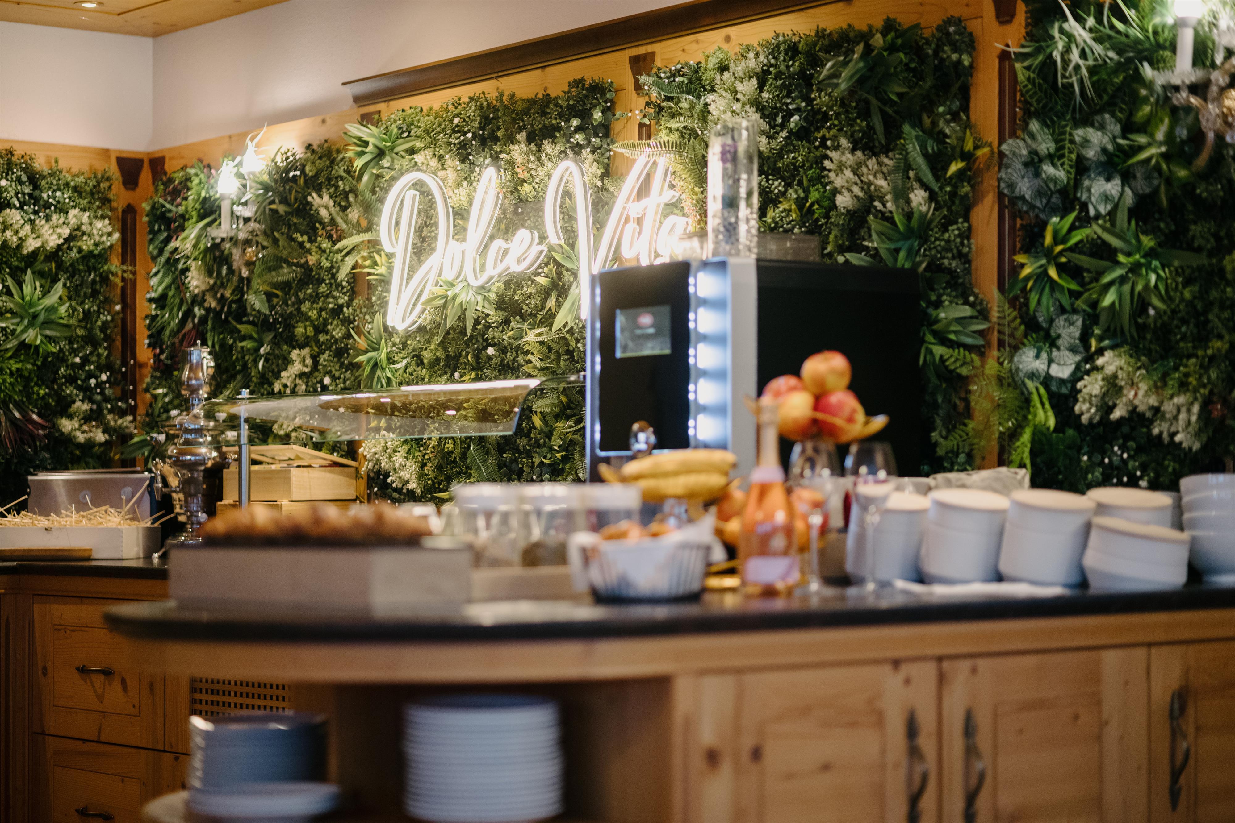 Buffet with fruit, dishes, and a green floral wall with neon sign in the background.