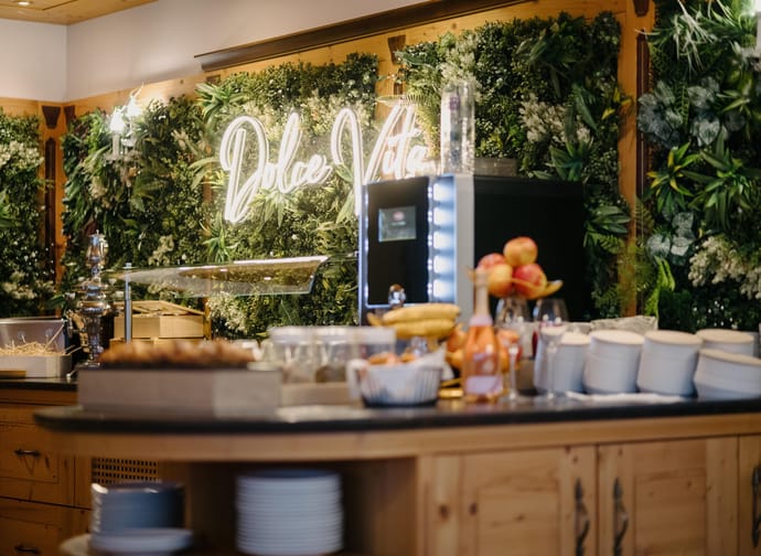 Buffet with fruit, dishes, and a green floral wall with neon sign in the background.