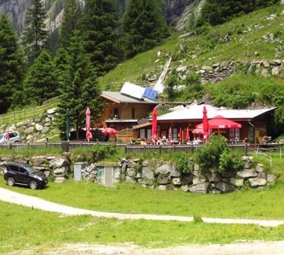 A mountain restaurant with red umbrellas, surrounded by green trees and rocks, with cars parked in front.
