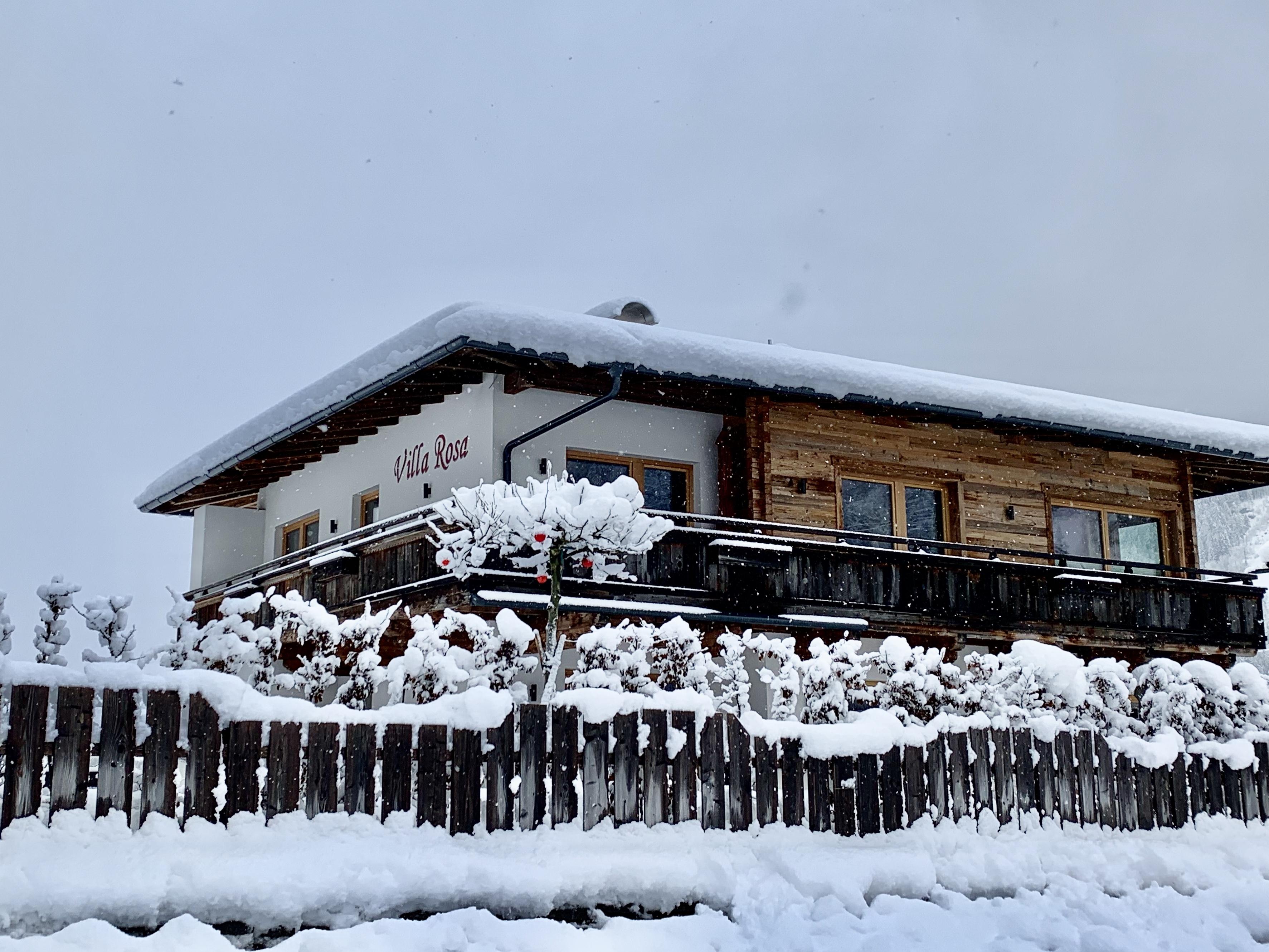 Snow-covered house with wooden facade and fence, surrounded by snow under a foggy sky.