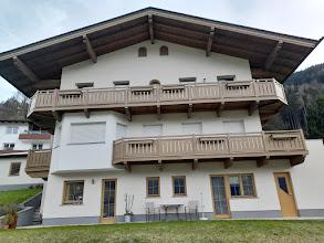 Multi-story house with white walls, wooden balconies, and a sloped roof in a mountainous area.