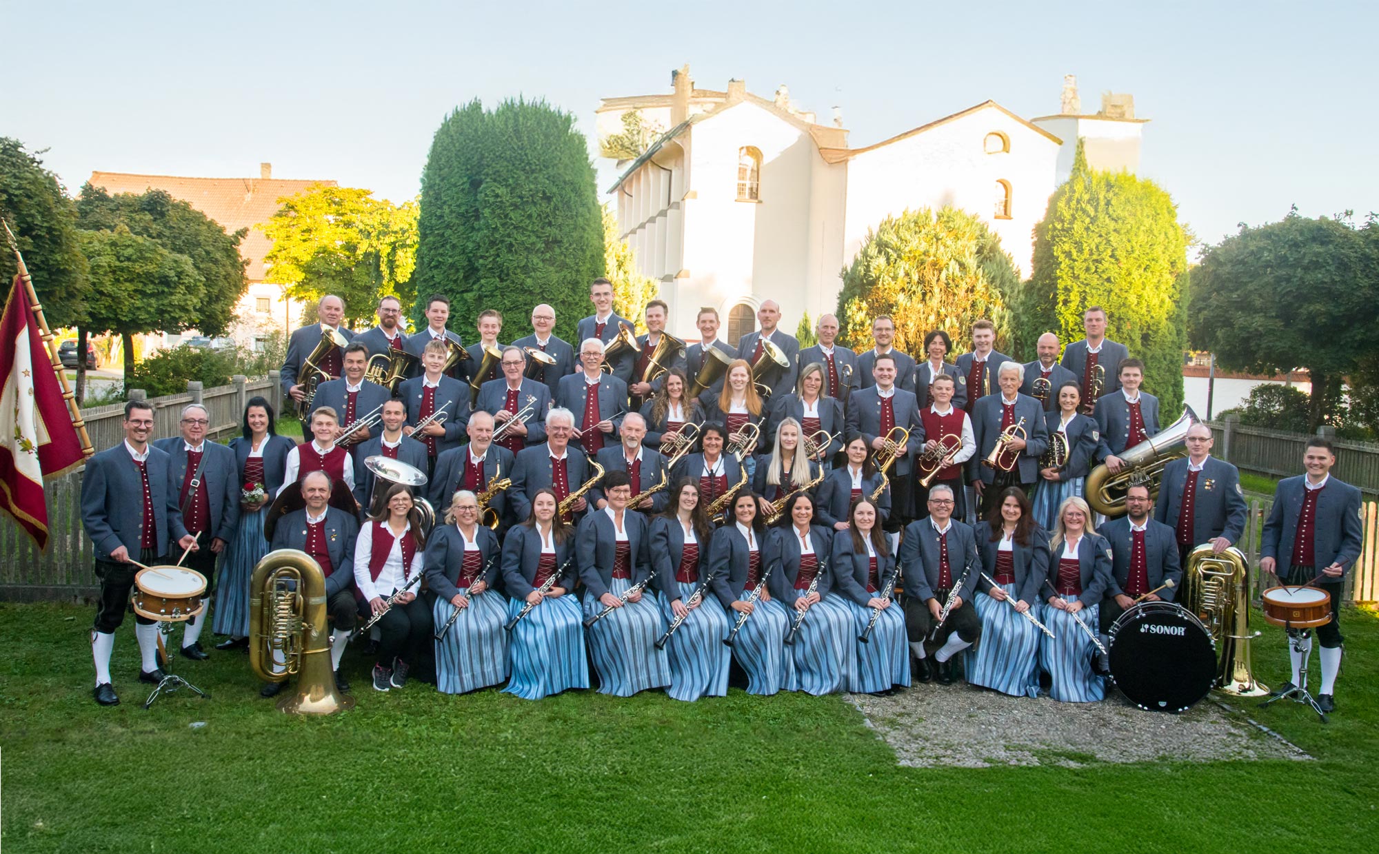 Das Bild zeigt eine große Blaskapelle in traditioneller Tracht, die vor einer Kirche für ein Gruppenfoto posiert, mit verschiedenen Blasmusikinstrumenten sowie Schlagzeug.