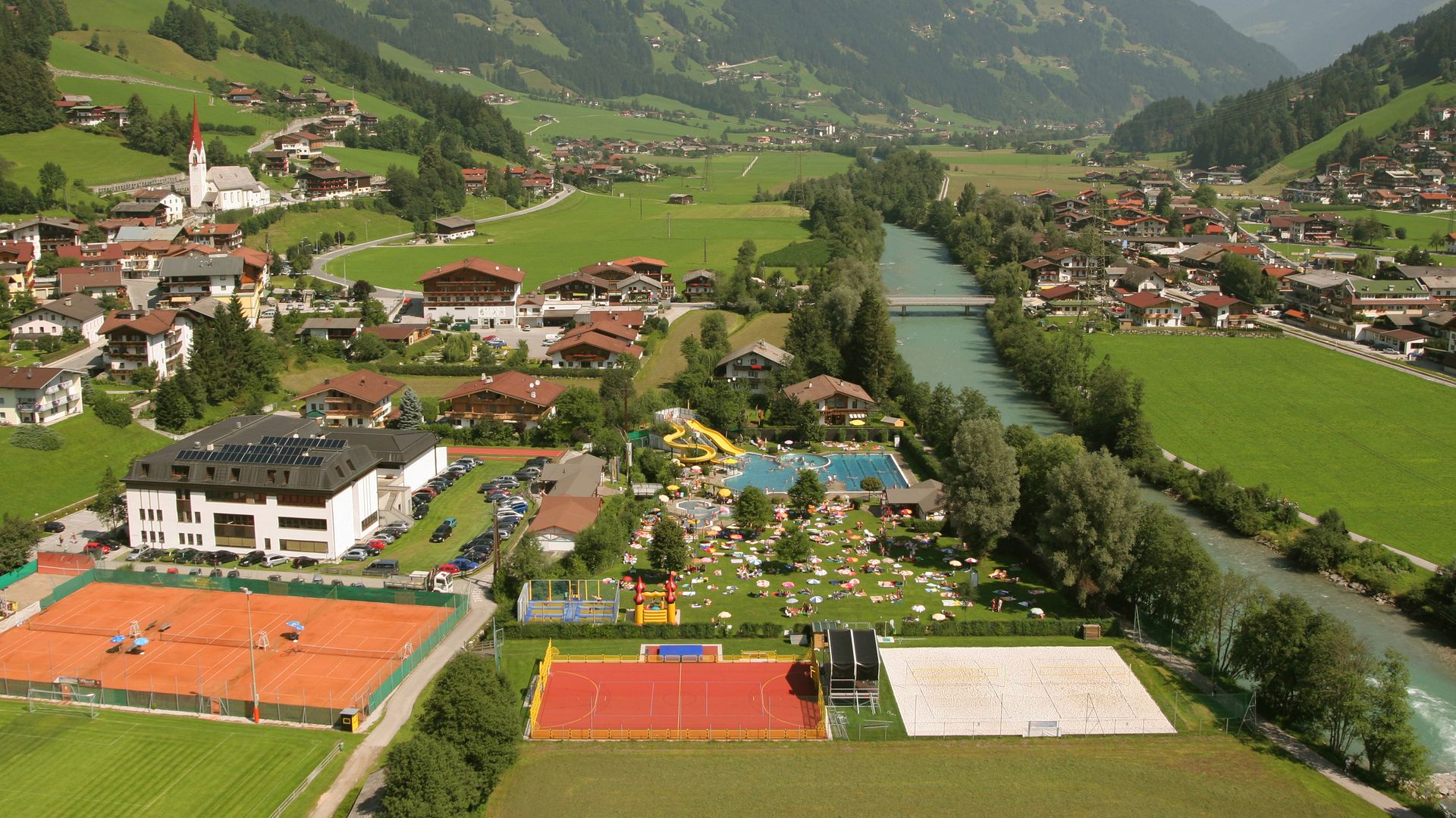Luftaufnahme der Sommerwelt Hippach mit Freibad, Sportplätzen und umliegender Berglandschaft im Zillertal an einem sonnigen Sommertag.