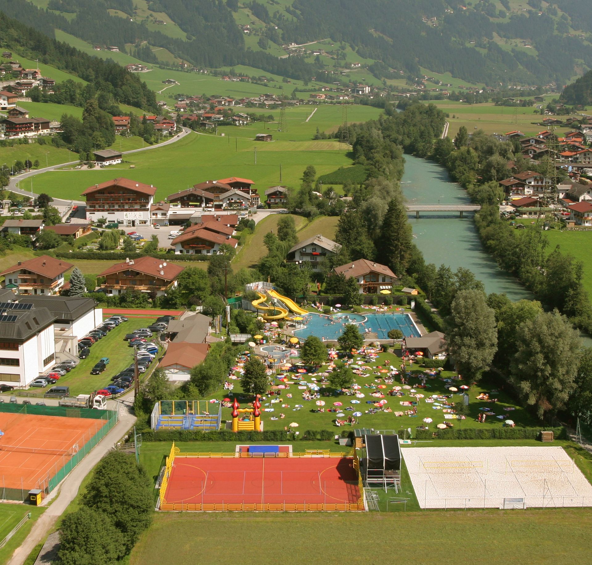 Luftaufnahme der Sommerwelt Hippach mit Freibad, Sportplätzen und umliegender Berglandschaft im Zillertal an einem sonnigen Sommertag.