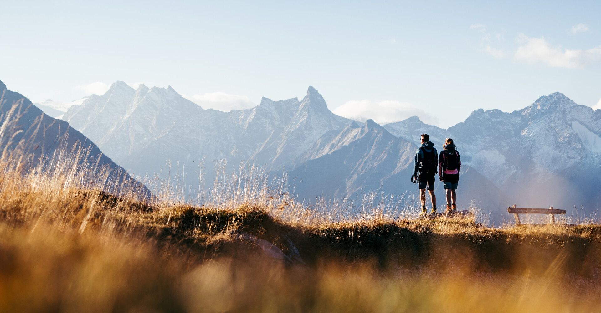 Two hikers enjoy the autumn view from Penkenjoch of the Zillertal Alps. Golden grass in the foreground and snow-capped peaks in the background.