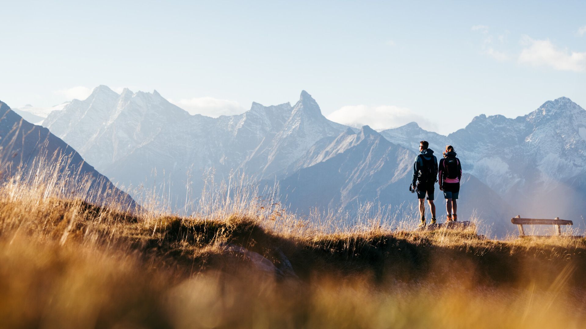 Two hikers enjoy the autumn view from Penkenjoch of the Zillertal Alps. Golden grass in the foreground and snow-capped peaks in the background.