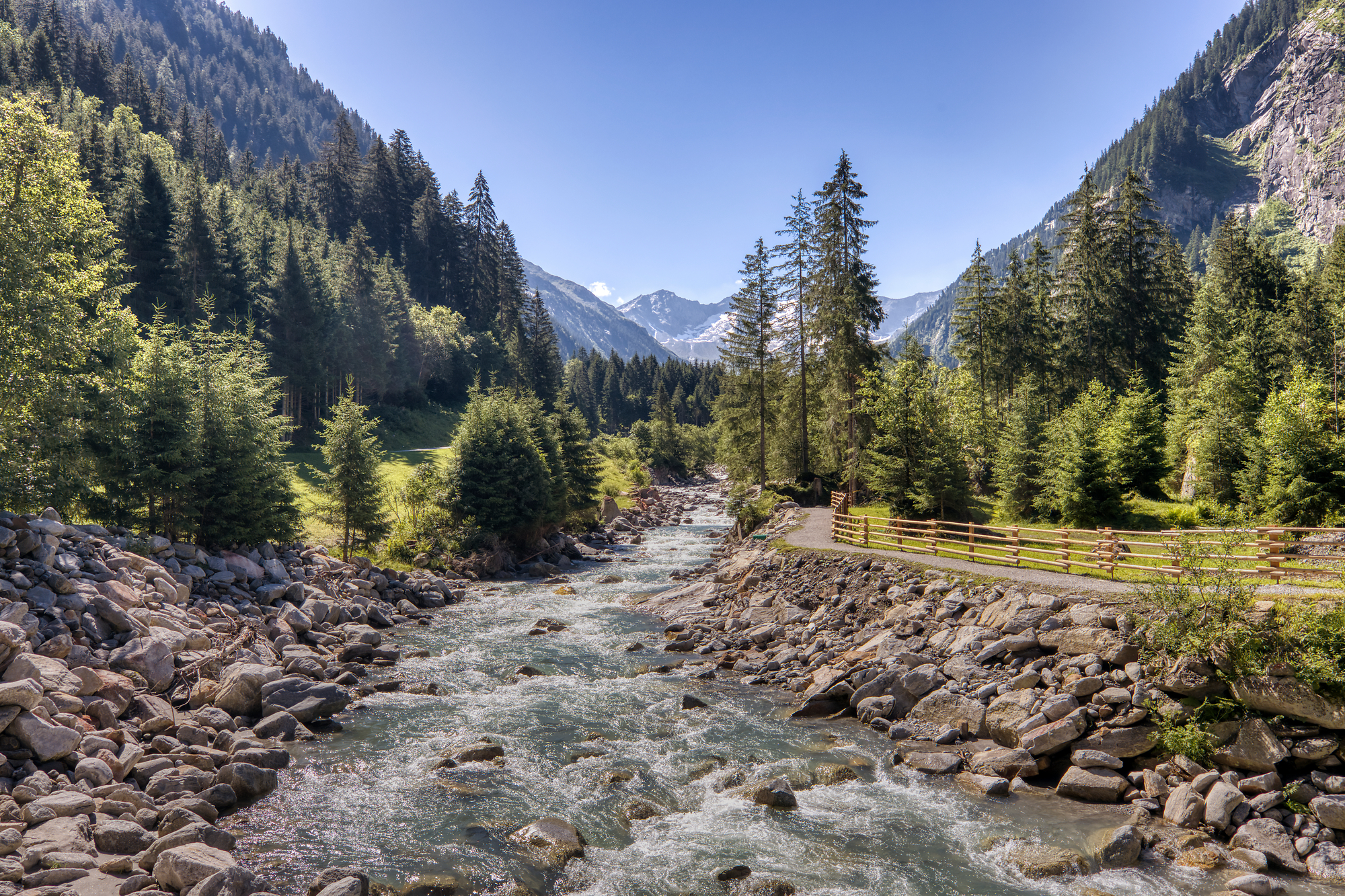 Ein Bach umgeben von Steinen, rechts ein Wanderweg und im Hintergrund Bäume und Berge