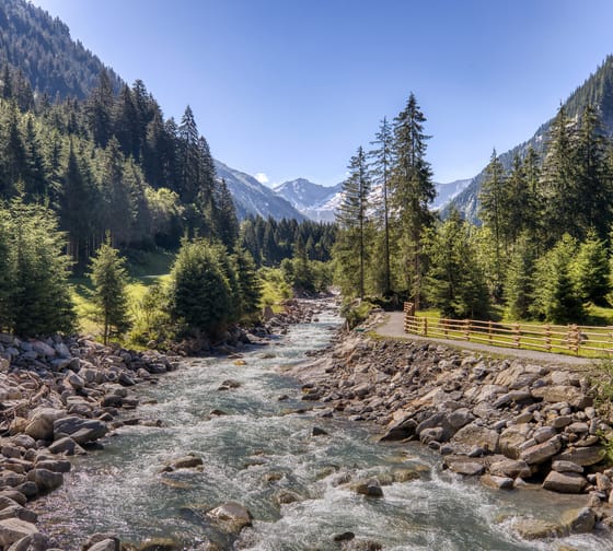Ein Bach umgeben von Steinen, rechts ein Wanderweg und im Hintergrund Bäume und Berge