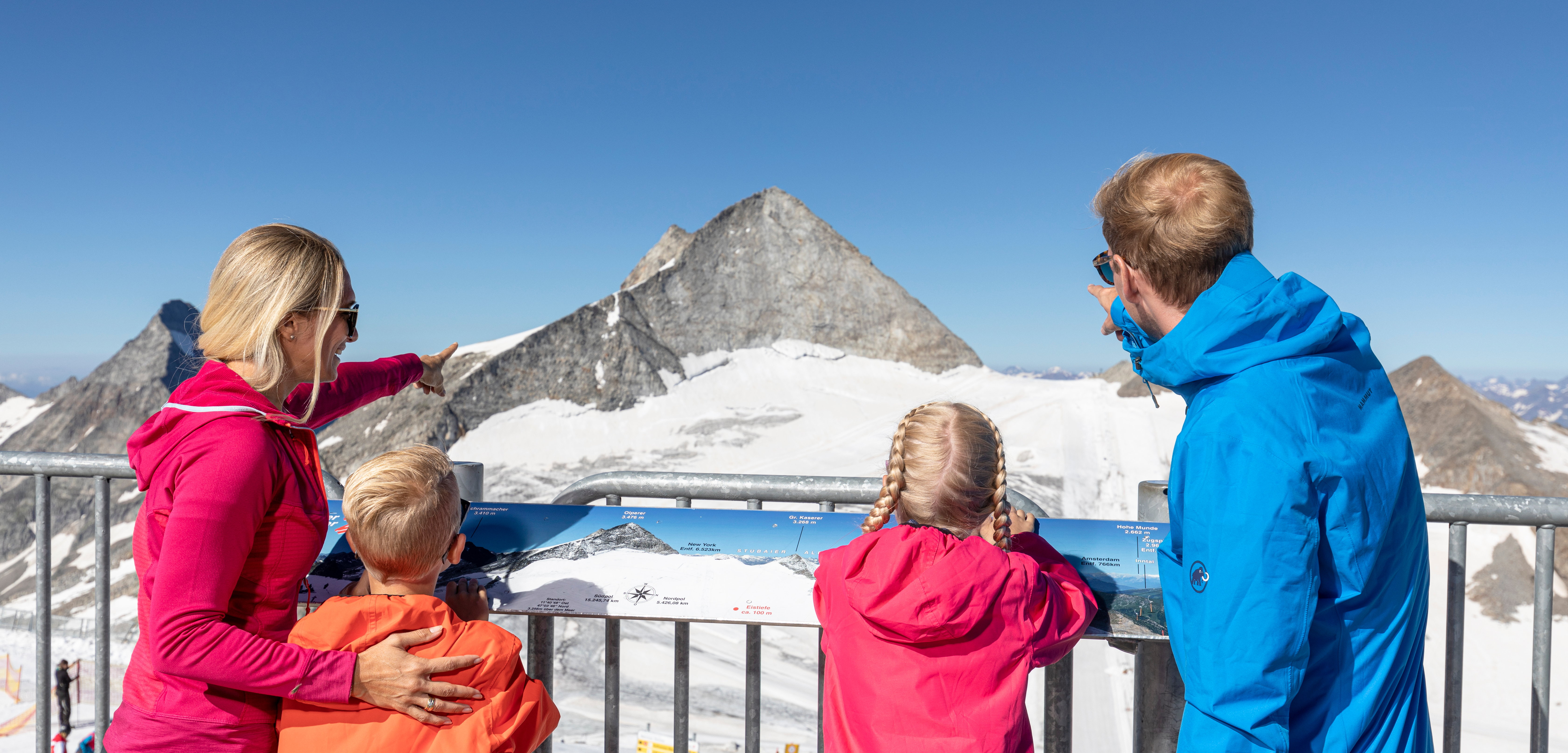Family at mountain summit, pointing at the peak, snowy mountains in background, sunny day, clear view.