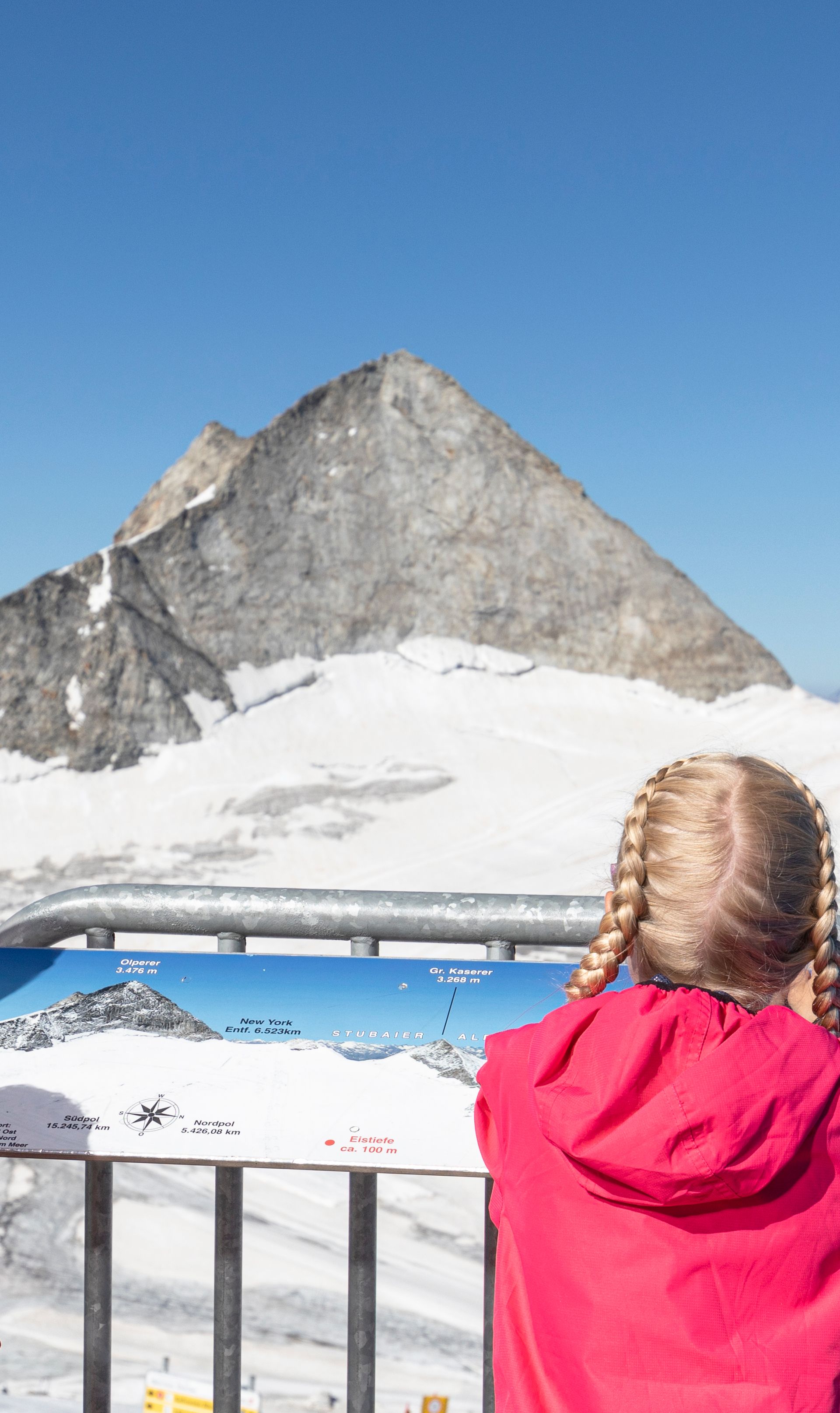 Family at mountain summit, pointing at the peak, snowy mountains in background, sunny day, clear view.