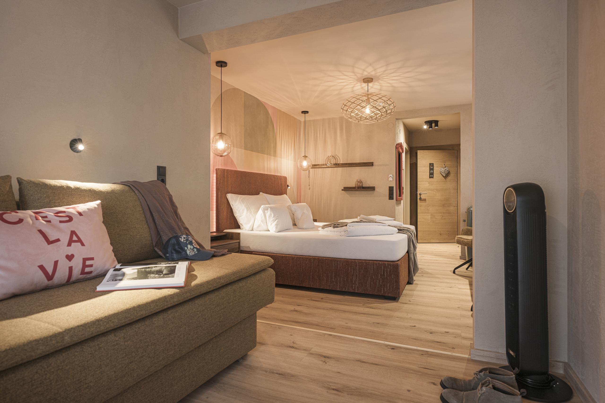 Gemütliches Schlafzimmer mit Bett, Sofa, Lampen und Holzwand, warme Beleuchtung, Schuhe am Boden.