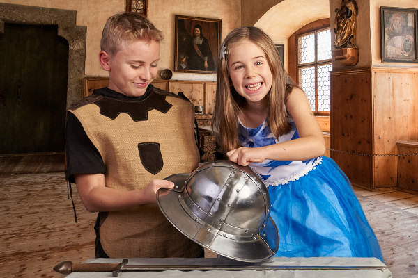 Two children in knight costumes laugh and look at a silver knight helmet in a historic room.