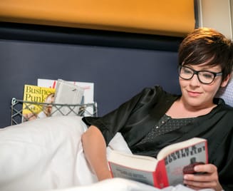 Frau mit Brille liest entspannt ein Buch im Nightjet-Schlafwagenabteil. Sie liegt bequem im Bett mit weißer Bettwäsche. Im Hintergrund sind Fenster und ein kleines Ablagenetz sichtbar.