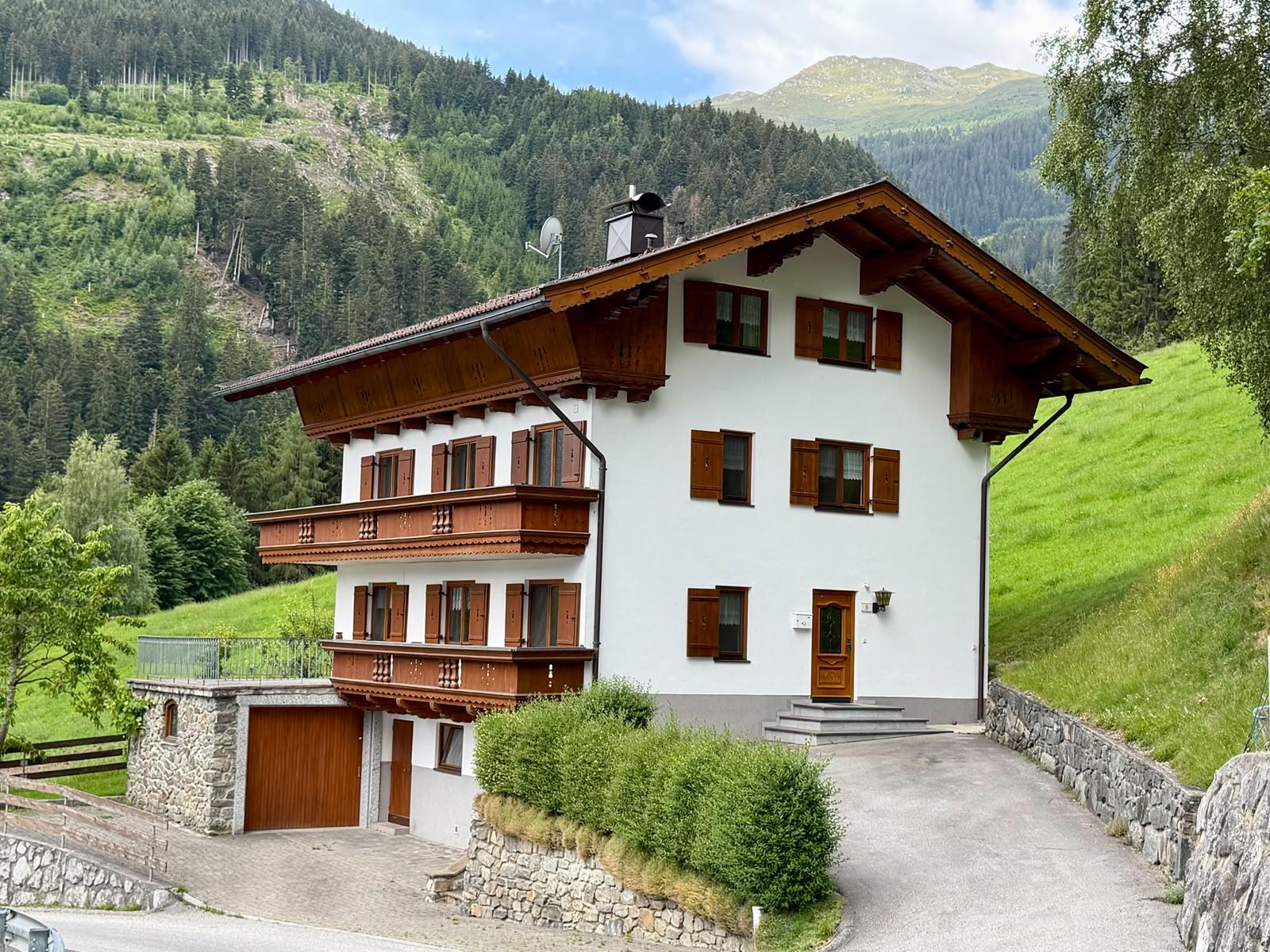 House with wooden balconies in a green, hilly landscape with mountains in the background.
