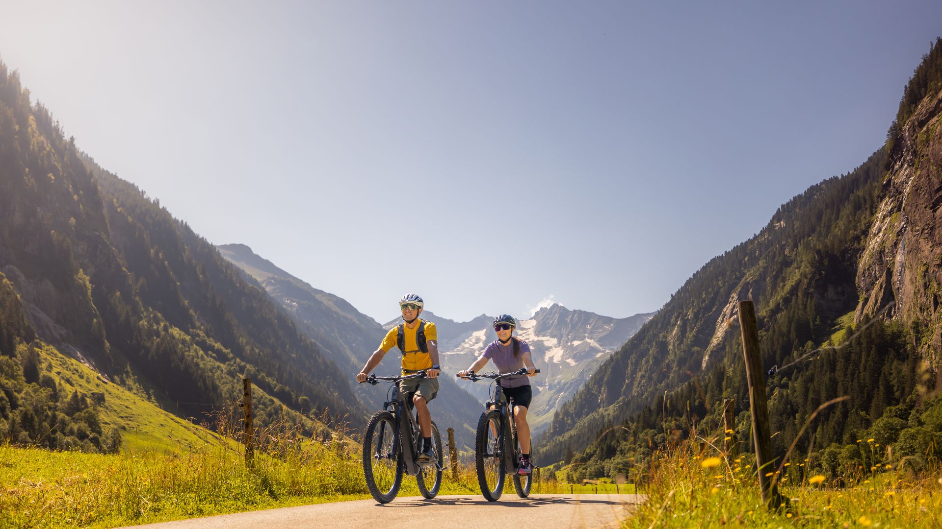 Moderate cycling tour near Mayrhofen-Hippach in the Zillertal: two people ride along a wide path through green meadows, surrounded by mountains under a blue sky.