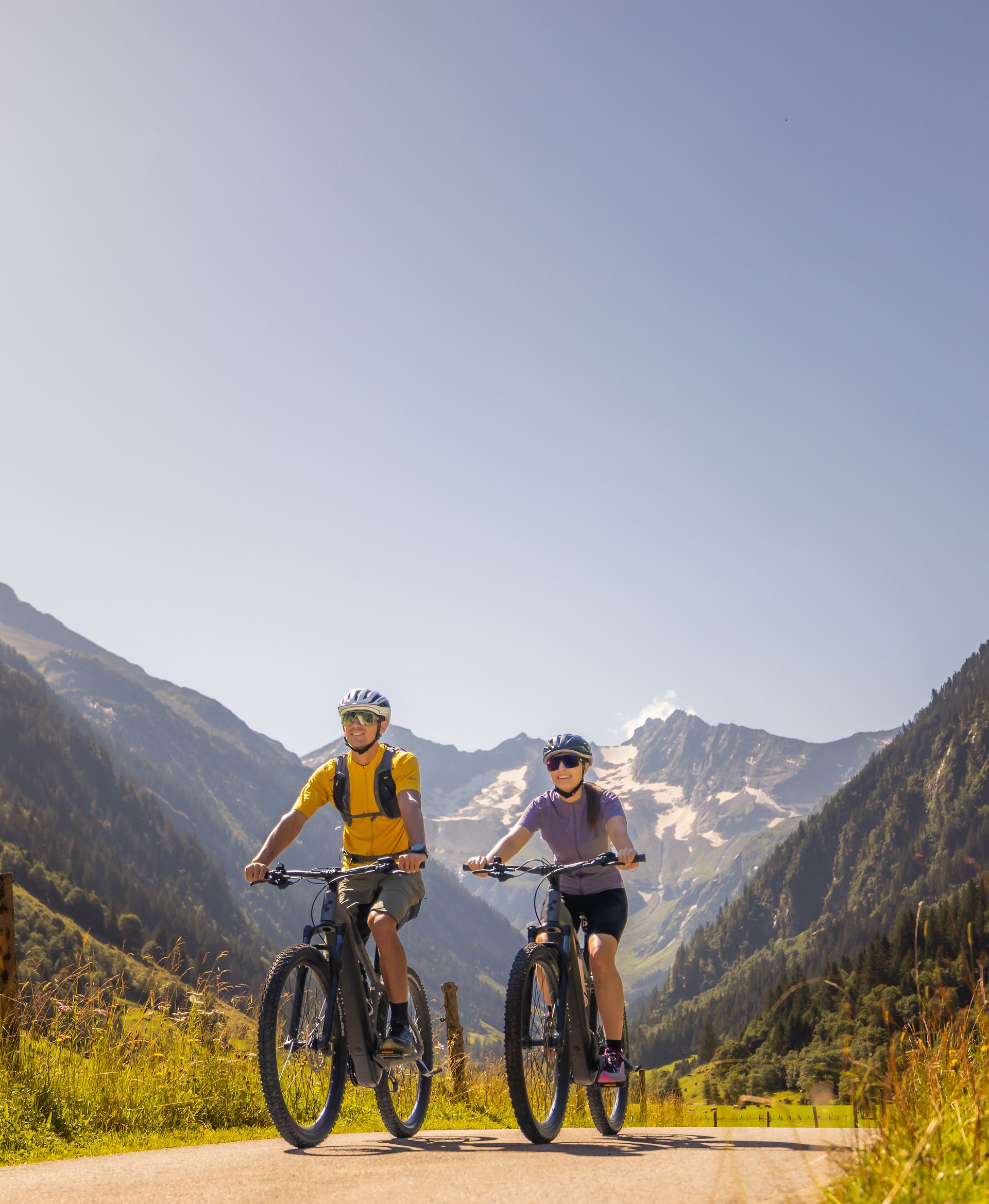Moderate cycling tour near Mayrhofen-Hippach in the Zillertal: two people ride along a wide path through green meadows, surrounded by mountains under a blue sky.