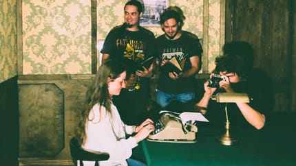 Das Bild zeigt eine Gruppe von vier Personen in einem Escape Room mit retro-inspirierter Einrichtung. Eine Frau sitzt an einem Tisch und tippt auf einer Schreibmaschine, während ein Mann durch eine Kamera schaut. Zwei Männer untersuchen ein Buch/Objekt.