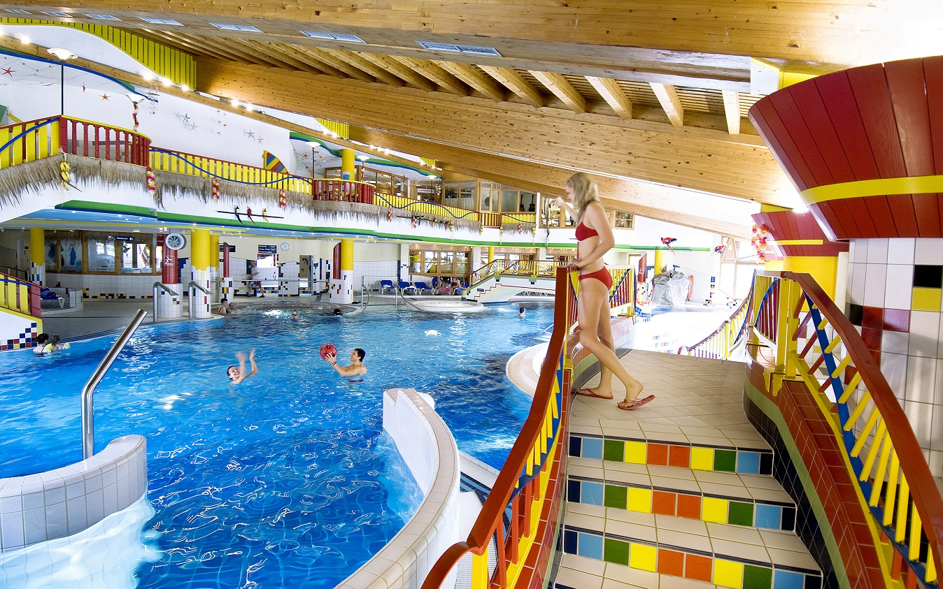 A bright, colorful indoor pool with a wooden ceiling. Children play in the water, a woman in a red bikini stands at the top of a colorful staircase. Slides, bridges, and railings in bold colors are visible in the background.