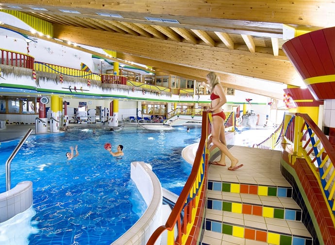 Ein helles, farbenfrohes Hallenbad mit Holzdecke. Kinder spielen im Wasser, eine Frau im roten Bikini steht oben an einer bunten Treppe. Im Hintergrund sind Rutschen, Brücken und Geländer in kräftigen Farben zu sehen.