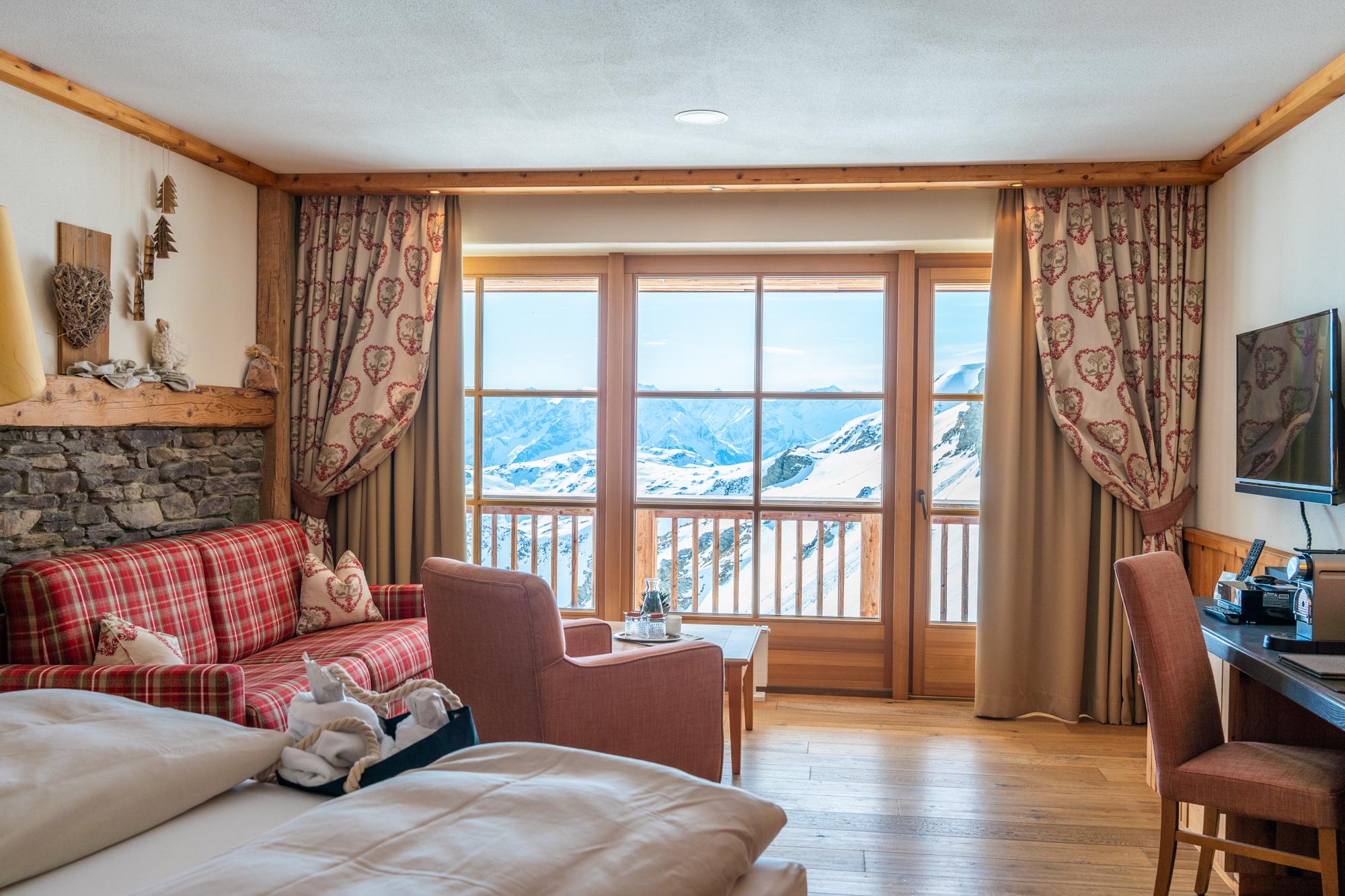 Gemütliches Hotelzimmer mit Blick auf verschneite Berge, Holzdekor, Sofa und Fernseher.