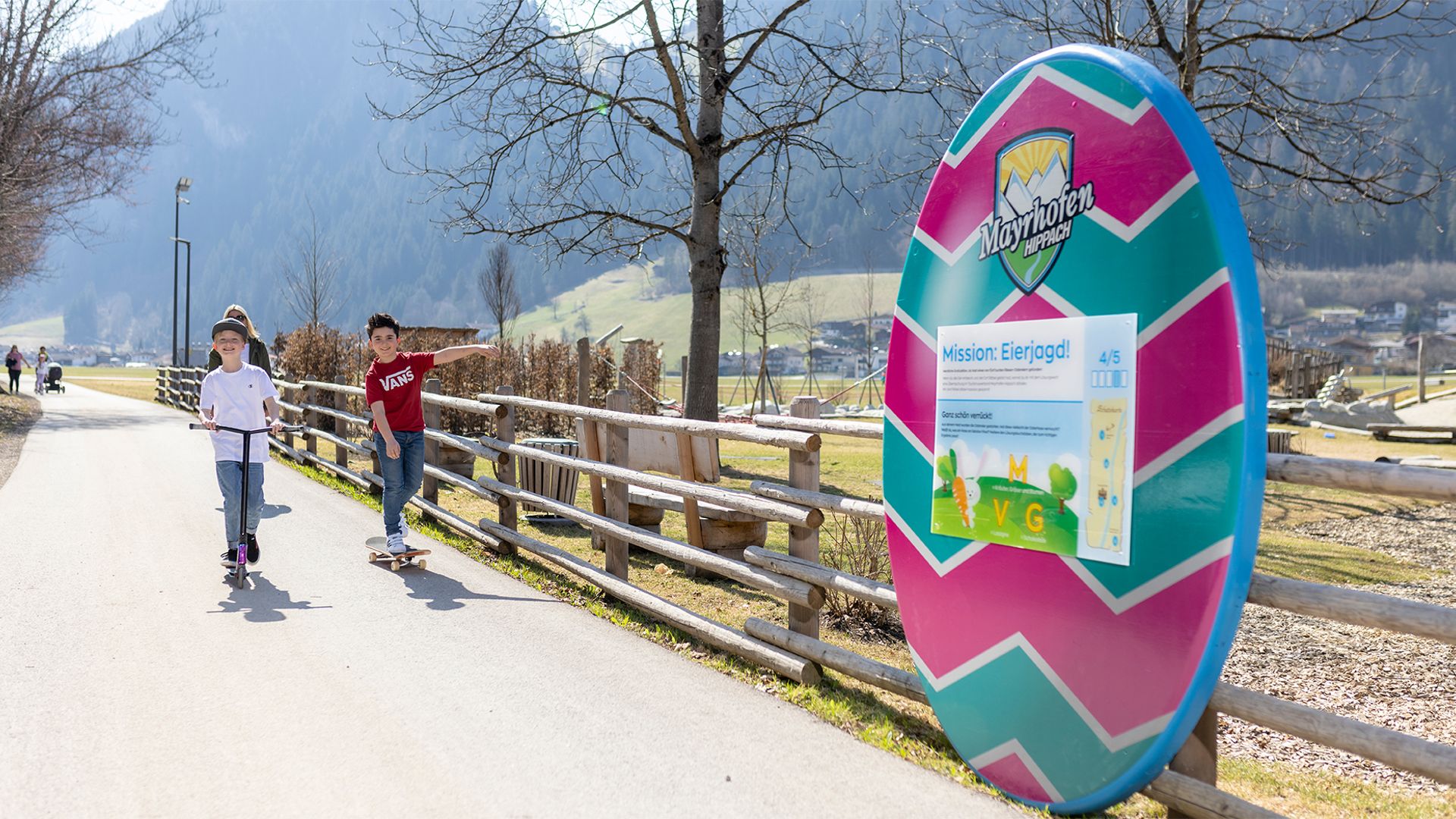 Zwei Kinder fahren auf einem Weg mit Roller und Skateboard. Daneben steht ein großes, buntes Osterei mit Infos zur „Mission Eierjagd“. Im Hintergrund sind Hügel, Bäume und Zäune zu sehen. Das Wetter ist sonnig und freundlich.