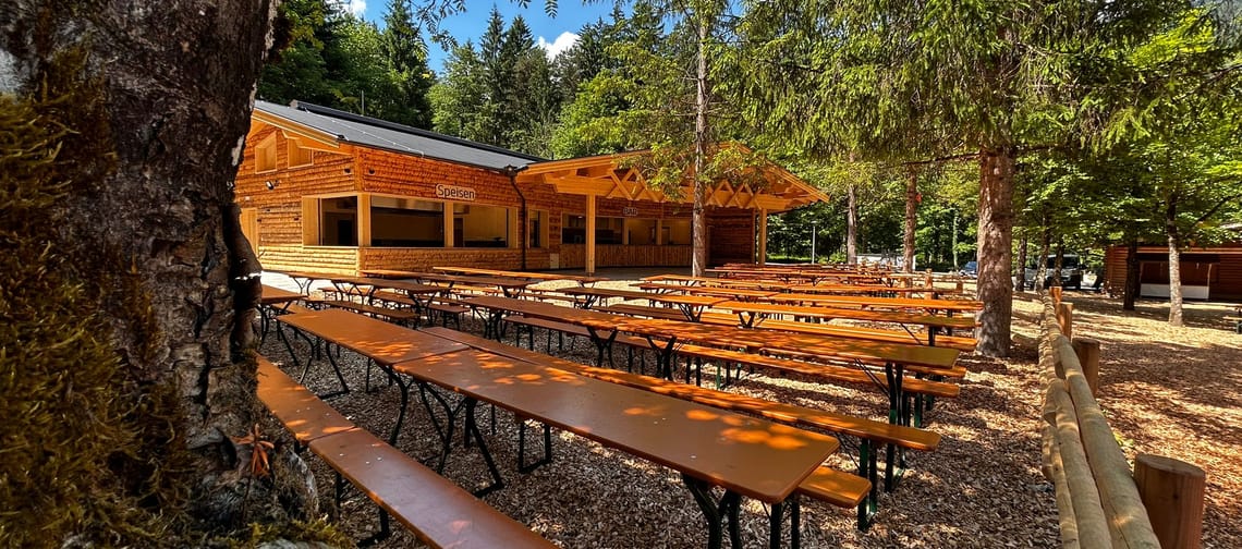 Im Bild sieht man ein Festgelände mit Festbankgarnituren im Wald. Der Boden ist mit Holzspänen ausgelegt, grüne Nadelbäume stehen neben den Bänken. Eine Holzblockhütte im Hintergrund.
