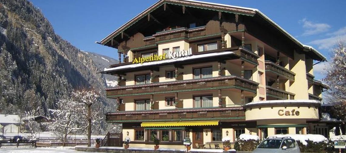 The image shows the Hotel Alpenhof Kristall from the outside in winter. Its light yellow facade is adorned with dark wooden balconies. In the background, snow-covered mountains rise into the clear blue sky