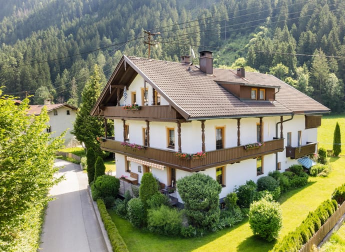 Bright house with balcony and flowers, surrounded by green trees and mountains in the background.