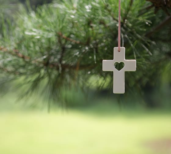 Das Bild zeigt ein Holzkreuz mit einem Herz in der Mitte, das an einem Tannenzweig hängt.