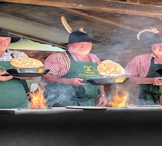 Das Bild zeigt 3 Männer in trachtiger Kleidung beim "Muaskochen" vor einem Herd.